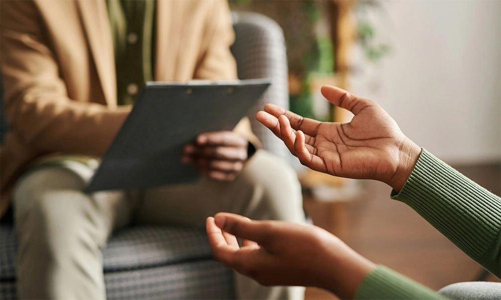Person holding documents in beige blazer, another person gesturing with open hands in green sweater.